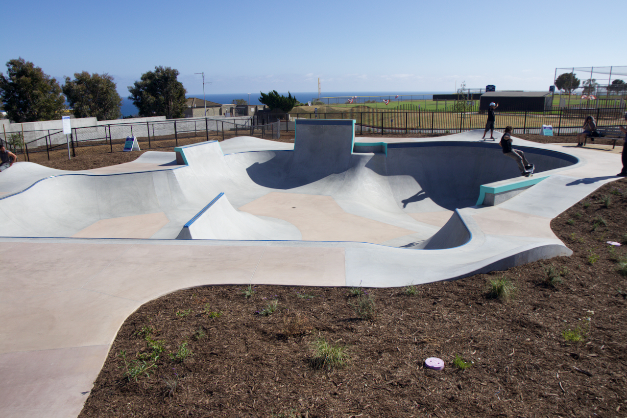Malibu's flow bowl from the shallow end, with a spine down the middle, a skater riding the far wall, and the ocean beyond the ball field.