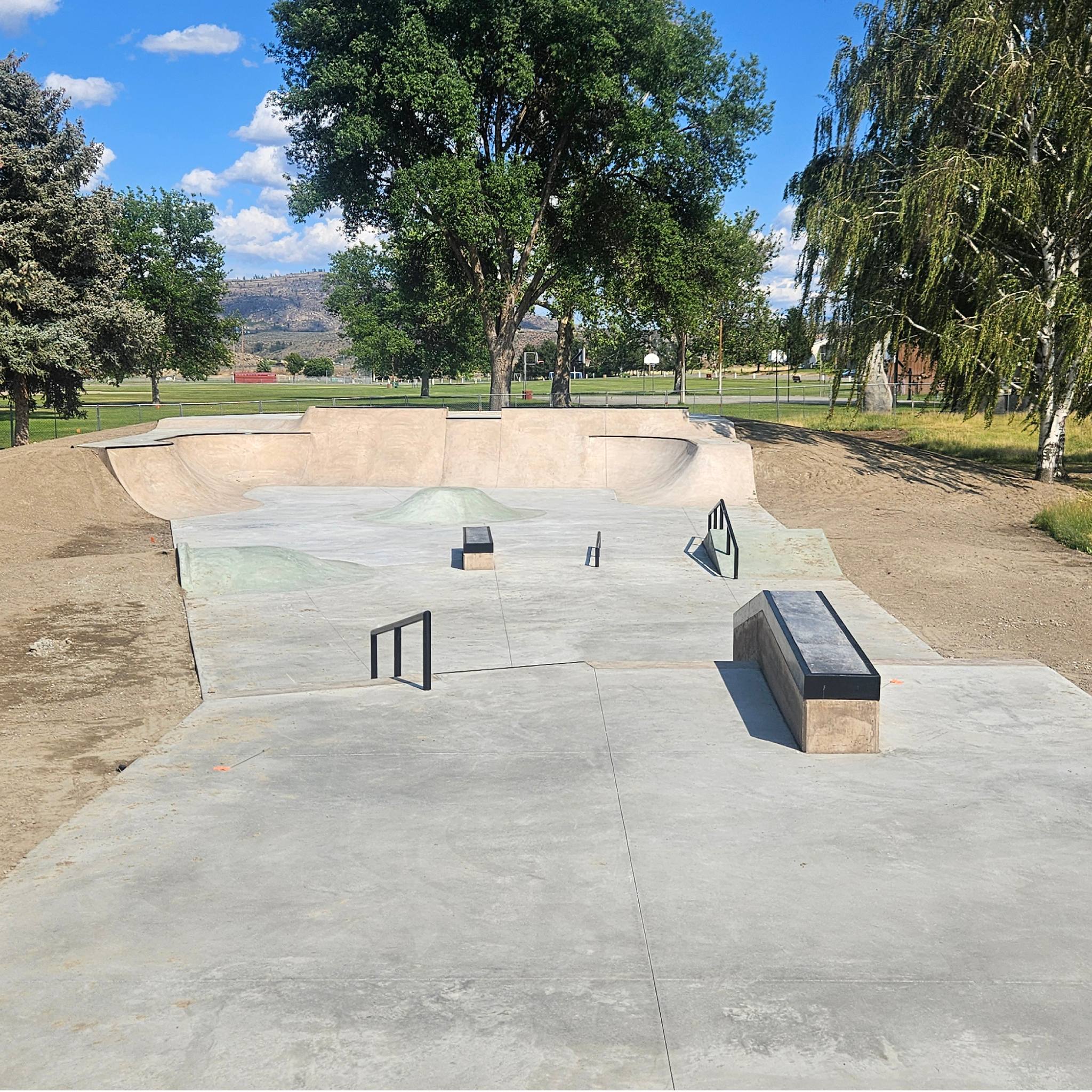 The street section at Omak's Eastside Park: a flat bar, a manual pad and a long hubba on the pad, with quarters and banks wrapping the far end under the trees.