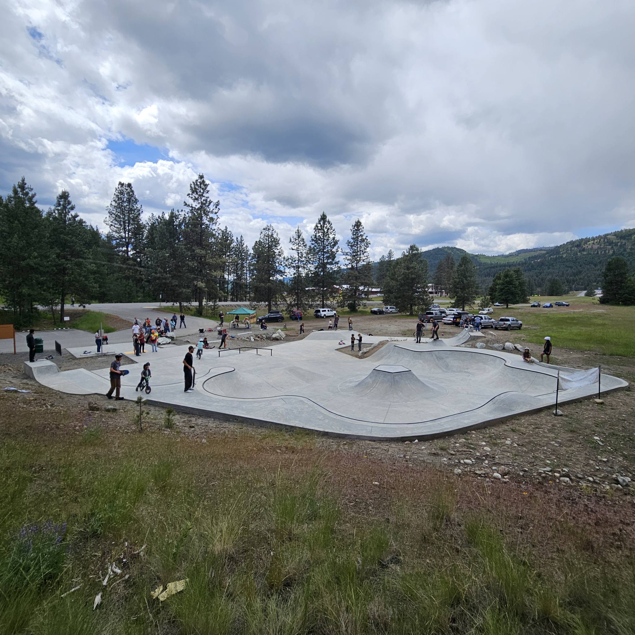 A crowd of riders and families on the decks at Republic Park, the lobed concrete bowl in the foreground and the pines and hills of Ferry County behind.