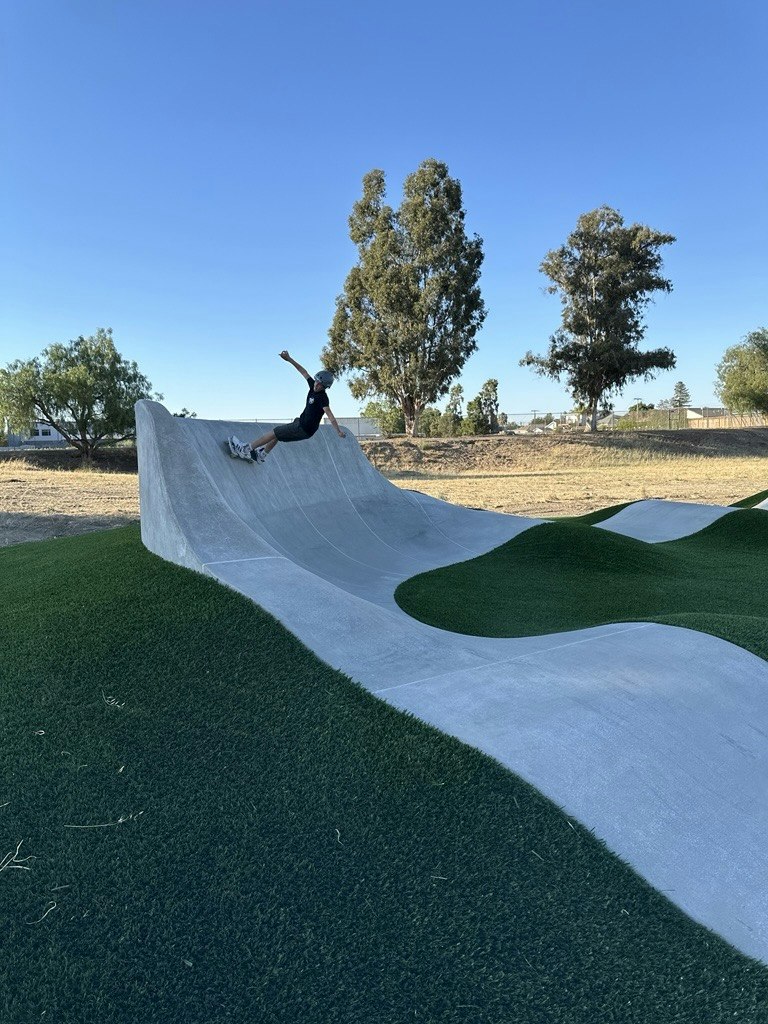 A skater carving the tall wall at the end of the Sunken Gardens pump track, turf hills rolling around it.